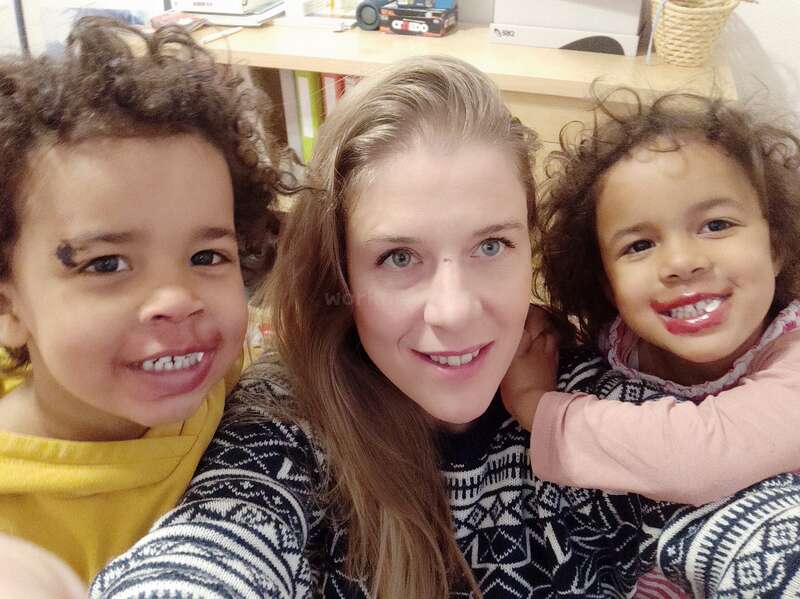 A imagem mostra uma mulher de cabelos longos posando para uma selfie com duas crianças, em frente a uma mesa com brinquedos e livros.
