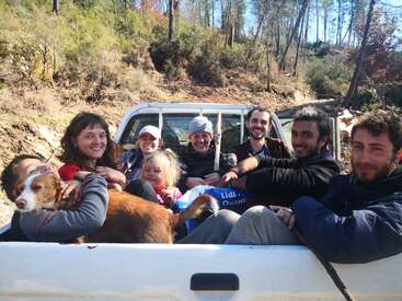 The image shows a group of people and a dog sitting in the back of a white pickup truck, parked on a dirt road in a wooded area.