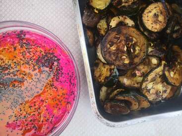 The image shows a bowl of pink dip with seeds on top and a baking tray of roasted vegetables on a tablecloth.