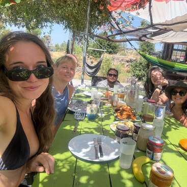 Six amis profitent ensemble d'un repas en plein air sous le soleil, assis à une table de pique-nique verte, entourés de nourriture, de bocaux et de boissons, à l'ombre des feuillages et sous des hamacs suspendus.
