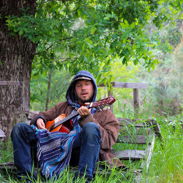 A man sits on a wooden bench in a grassy area, playing a guitar, wearing a brown jacket and blue jeans, with a blue and white patterned blanket nearby.