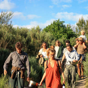 A group of people, including a child, walk along a dirt path through a grassy area, carrying various items, with trees and a blue sky in the background.