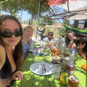 Seis amigos disfrutan juntos de una comida al aire libre bajo el sol, sentados en una mesa de picnic verde, rodeados de comida, tarros y bebidas, bajo la sombra de las hojas y hamacas colgantes.