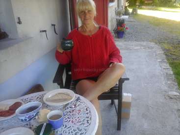 La imagen muestra a una mujer sentada en una silla al aire libre, sosteniendo una taza, con una mesa delante en la que hay una comida y una taza de té.