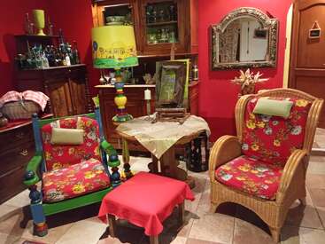 Esta acogedora habitación cuenta con sillones con estampados florales de colores, una lámpara original, paredes rojas, muebles de madera, un espejo decorativo, botellas, cestas y una mesita con un mantel.