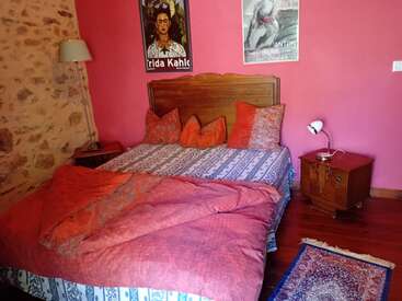 Un dormitorio acogedor con paredes rosas, una cama grande con ropa de cama naranja, cabecero de madera, mesitas de noche, lámparas, pósters, una pequeña alfombra y una pared decorativa de piedra.
