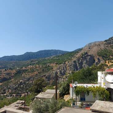 The image depicts a serene mountainous landscape with a small village at its base, featuring white buildings, lush trees, and a clear blue sky.