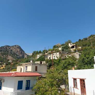 The image depicts a serene hillside village, featuring white buildings with red roofs, lush greenery, and a majestic mountain in the background under a clear blue sky.