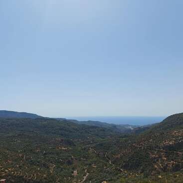 The image depicts a serene mountainous landscape with lush greenery, featuring a winding road and a distant body of water, set against a clear blue sky.