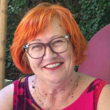The image depicts a woman with short, bright orange hair, wearing a pink top and glasses, posing for a selfie in front of a leafy background.