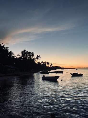 Boote treiben bei Sonnenuntergang sanft auf dem ruhigen Wasser, Silhouetten von Palmen säumen das Ufer und der Himmel wechselt von Orange zu tiefblauem Dämmerlicht.