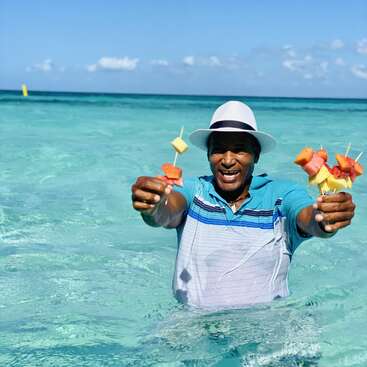 The image depicts a man standing in shallow water, wearing a white hat and blue shirt, holding two fruit skewers in each hand, with a serene ocean and sky background.