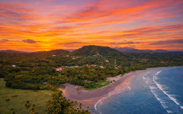 A vibrant sunset fills the sky with orange and purple hues above a lush tropical coastline, palm trees, gentle waves, and a small village nestled by green hills.