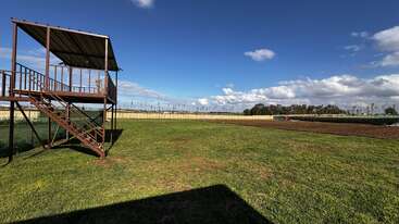 A wooden platform with stairs stands on a large grassy field, surrounded by fences. The sky is blue with scattered clouds, creating a peaceful rural scene.