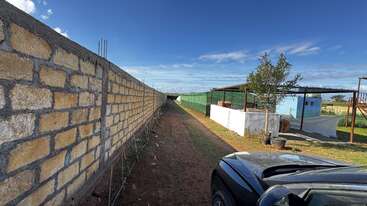 A brick wall runs alongside a dirt path. On the right, there’s a parked car, a tree, fenced area and buildings under a bright blue sky.