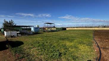 This image shows a wide, open grassy field under a blue sky with scattered clouds. There are sheds, a water tank, and farming equipment visible.