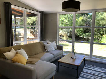 This bright living room features large windows, a comfortable grey couch with pillows, a wooden coffee table, a striped rug, and overlooks a lush green garden.