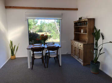 A cozy dining room features a wooden table set for four, a large window with a garden view, indoor plants, and a rustic wooden hutch.