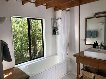 This is a bright bathroom with a bathtub by a large window, wooden ceiling beams, a sink on a wooden counter, and greenery visible outside.