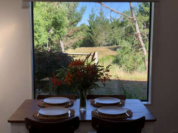 A dining table set for four sits by a large window, offering a beautiful view of a sunny, green outdoor landscape and tall trees.