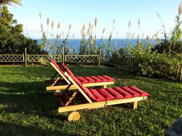 L'image représente deux chaises longues en bois avec des coussins rouges sur une pelouse luxuriante, surplombant un plan d'eau serein sous un ciel bleu clair.