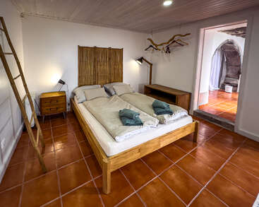 Cette chambre à coucher confortable se compose d'un lit en bois avec une literie beige, de carreaux de sol en terre cuite, d'un mobilier minimaliste, d'une tête de lit en bambou, d'une échelle rustique et d'une atmosphère bien éclairée et accueillante.