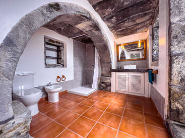 Cette salle de bains présente des arches en pierre rustiques, des carreaux de sol en terre cuite, une douche moderne, des toilettes et un bidet. Elle allie une architecture traditionnelle à des installations contemporaines et à un éclairage chaleureux.