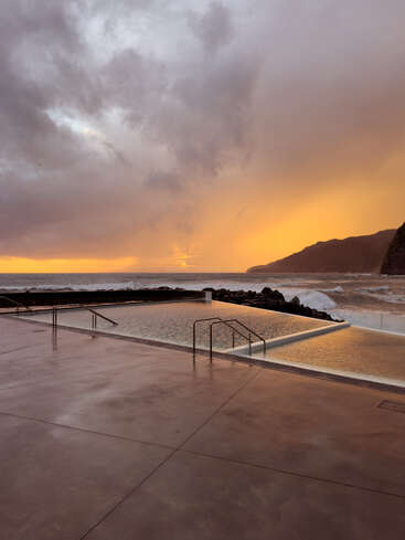 Un coucher de soleil serein jette une lumière dorée sur une piscine paisible au bord de l'océan, avec des nuages spectaculaires au-dessus de la tête et des falaises lointaines qui se découpent sur le ciel rougeoyant.