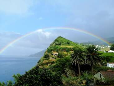 Un arc-en-ciel vibrant s'étend sur un village verdoyant au sommet d'une colline avec des palmiers, surplombant un océan calme, sous un ciel bleu partiellement nuageux. Un paysage paisible.