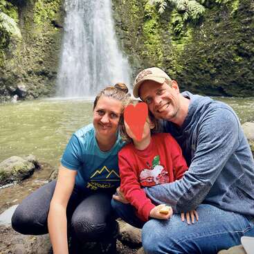 Une famille souriante pose ensemble devant une belle cascade entourée d'un feuillage vert luxuriant. Le visage de l'enfant est recouvert d'un cœur rouge dessiné.