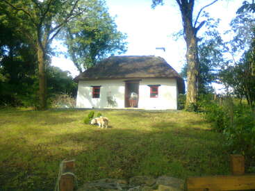 Une charmante maison blanche au toit de chaume est entourée d'arbres. Un petit chien joue sur la pelouse ensoleillée. Atmosphère paisible.
