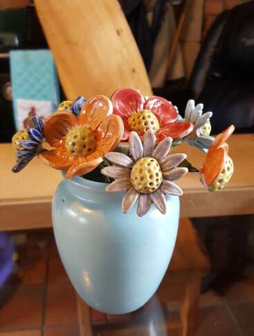 Un petit vase bleu contient des fleurs colorées en céramique avec un centre jaune, joliment disposées. L'arrière-plan semble être un intérieur confortable et chaudement éclairé.