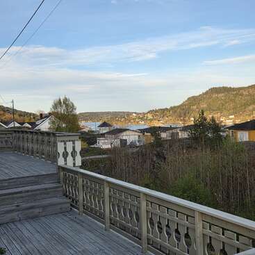 A wooden deck overlooks a peaceful neighborhood with houses and greenery. In the distance, there are hills, trees, and a glimpse of water under a blue sky.