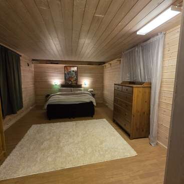 This cozy bedroom features wooden walls and ceiling, a large bed, two nightstands, dresser, artwork, gray curtains, and a fluffy white rug on wooden floor.