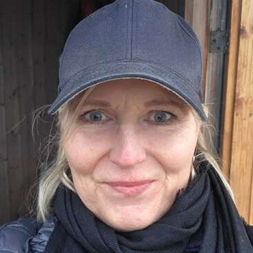 Eine Frau mit blonden Haaren und blauen Augen lächelt sanft. Sie trägt eine schwarze Baseballkappe und einen schwarzen Schal und steht vor einem hölzernen Hintergrund.