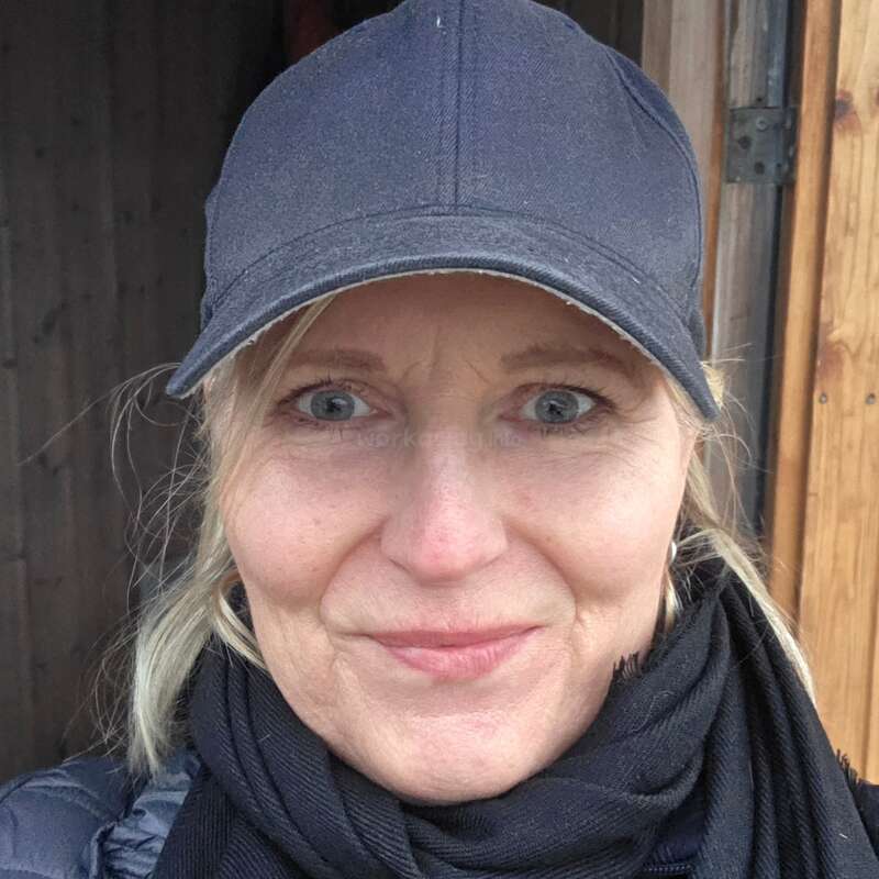 A woman with blonde hair and blue eyes smiles softly. She wears a black baseball cap, a black scarf, and stands in front of a wooden background.
