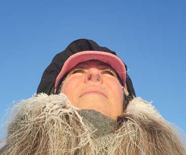 A person faces upward, bundled in winter clothing with frosted hair and scarf, under a clear blue sky, showing the effects of extreme cold weather.