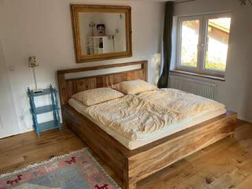 Un acogedor dormitorio con una cama de madera, ropa de cama con motivos amarillos, una mesita auxiliar azul, un espejo grande, una ventana, suelo de madera noble y un reflejo divertido.
