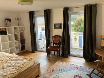 Se trata de un acogedor dormitorio con suelos de madera, una cama, estanterías blancas con ropa de cama, una silla clásica de madera, dos ventanas grandes con cortinas oscuras y una alfombra de colores.