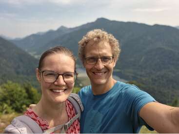 Una pareja sonriente, ambos con gafas, se hace un selfi al aire libre. Detrás de ellos se ven unas hermosas montañas verdes y un cielo brumoso, lo que sugiere una excursión por la montaña.
