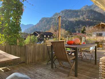 Una acogedora terraza de madera con una mesa, sillas y calabazas ofrece vistas a encantadoras casas y a unas montañas frondosas y bañadas por el sol bajo un cielo azul y despejado. Un ambiente sereno y acogedor.