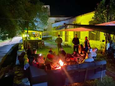 A group of people gathers around a cozy campfire at night, chatting and relaxing outdoors beside a bright, yellow-lit building, creating a warm, festive atmosphere.