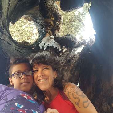 L'image montre une femme et un garçon posant pour un selfie devant un grand arbre troué, entouré de branches et de feuilles.