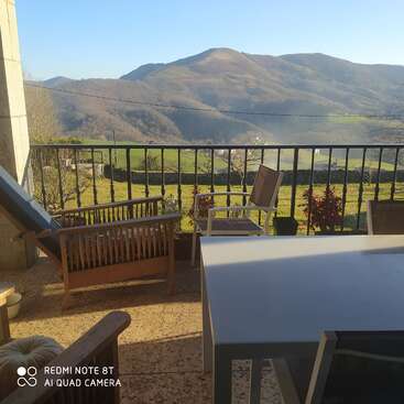 L'image représente un balcon serein avec une table et des chaises, offrant une vue pittoresque sur des collines et des montagnes ondulantes sous un ciel bleu clair par une journée ensoleillée.