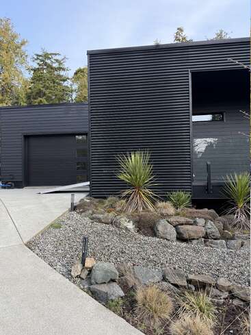 Uma casa preta moderna com revestimento horizontal, cercada por uma paisagem minimalista com pedras, cascalho e plantas desérticas pontiagudas. A entrada de automóveis de concreto se curva ao longo da estrutura.
