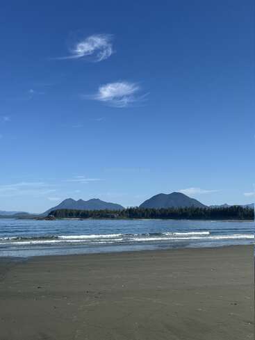 Uma praia de areia serena com ondas suaves, oceano azul calmo, montanhas arborizadas distantes e um céu claro adornado com nuvens finas, criando uma paisagem pacífica e natural.