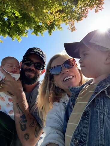 Una feliz familia de cuatro miembros posa para un selfie al aire libre, con la luz del sol brillando a través de las hojas verdes. Los padres sonríen con sus dos hijos pequeños en brazos.