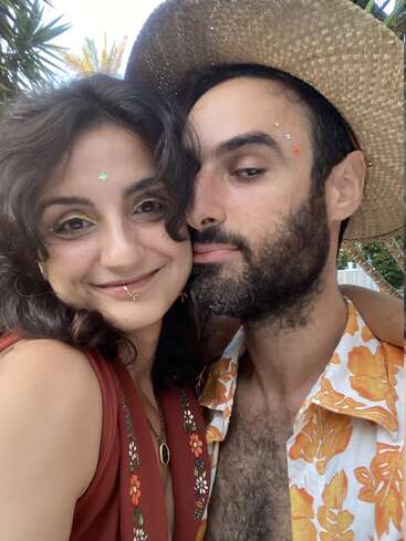 Um casal feliz posa junto, ambos sorrindo. A mulher usa adesivos faciais decorativos e um vestido floral, enquanto o homem usa um chapéu de palha e uma camisa floral.