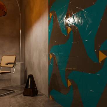 This image shows a modern, minimalist room with a tan chair, angular wooden object, and a colorful geometric mural featuring blue, brown, and yellow shapes on the wall.