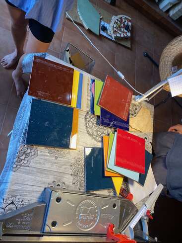 The image shows colorful tiles arranged on a patterned tablecloth, a tile cutter, and two people standing nearby in a room with brown tiled floors.
